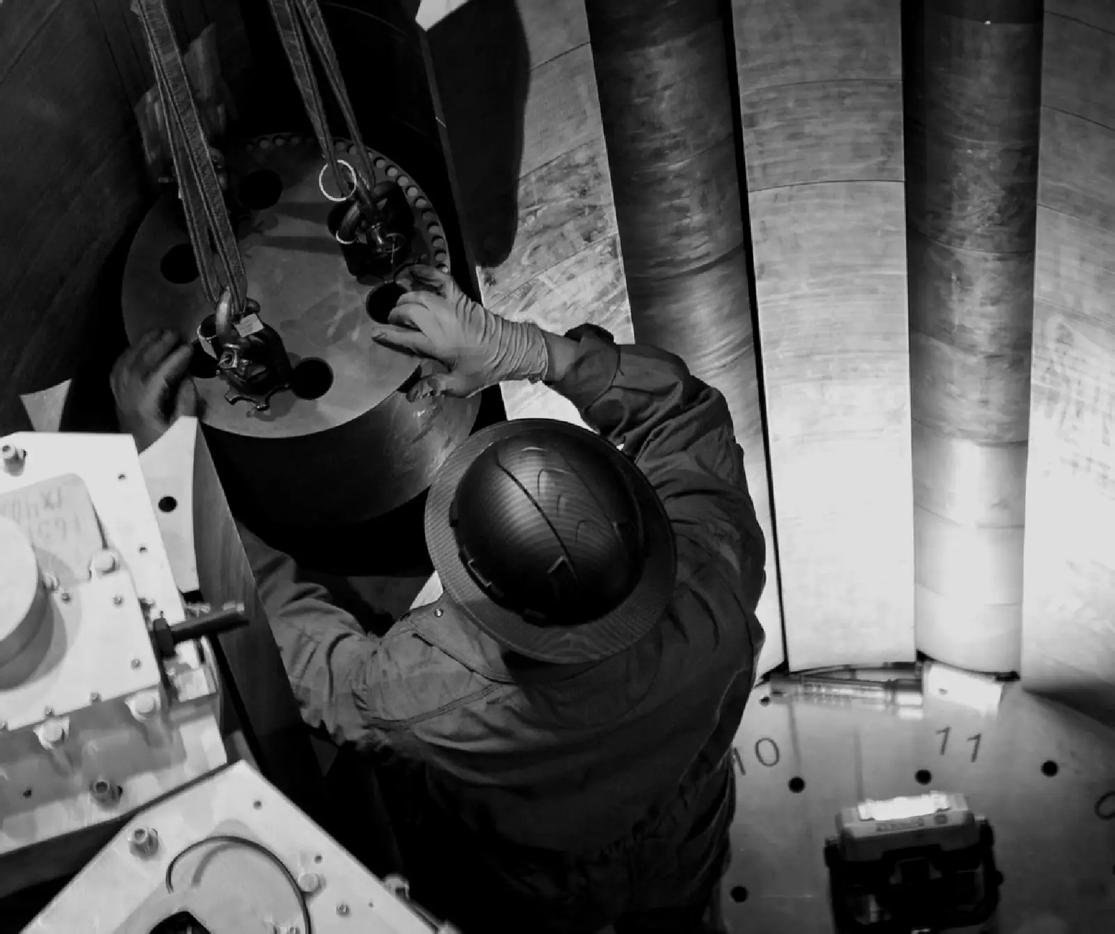 Two technicians in hard hats guide a cylindrical core component on lifting straps between tall graphite columns inside the high bay.